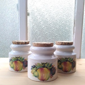 Peut inclure: Trois pots en céramique blanche avec des couvercles en liège et des illustrations de fruits. Chaque pot présente un motif de fruit différent, notamment des pommes, des prunes et des baies. Les pots sont parfaits pour ranger les épices, le thé ou d'autres produits de cuisine essentiels.