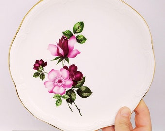 May include: White ceramic plate with a gold rim and a floral design featuring pink and purple roses.
