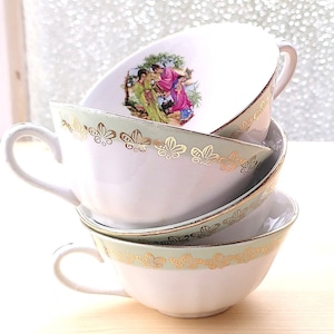 Peut inclure: Une pile de trois tasses à thé blanches avec des bordures dorées et vertes. La tasse du haut présente un motif floral avec un couple au centre.