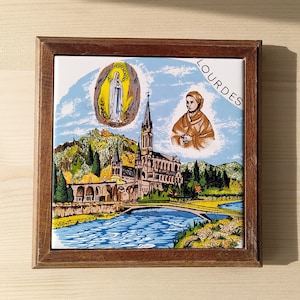 May include: A framed tile with a colorful illustration of the Sanctuary of Our Lady of Lourdes in Lourdes, France. The tile features a depiction of the Virgin Mary, a church, and a river. The tile is surrounded by a brown wooden frame and has the word "Lourdes" printed in the upper right corner.