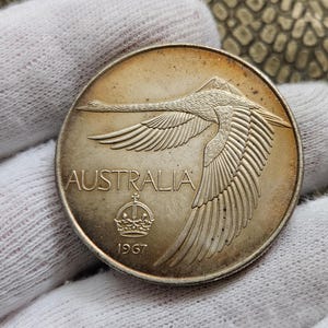 Könnte beinhalten: Silberne australische Münze mit einem stilisierten schwarzen Schwan im Flug, darunter die Worte "AUSTRALIA" und "1967".