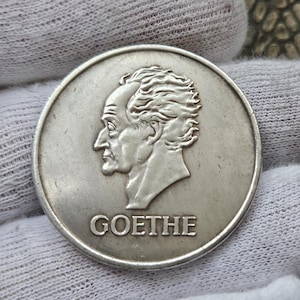 Puede incluir: Moneda de plata con un perfil de Johann Wolfgang von Goethe, escritor, poeta y filósofo alemán. La moneda tiene la palabra "GOETHE" inscrita debajo del perfil.