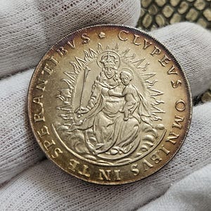 Op de afbeelding: Een ronde metalen munt met een reliëf van een gekroonde figuur die een kind vasthoudt, omringd door een zonnestraalpatroon. De Latijnse tekst "CLYPEVS OMNIBVS IN TE SPERANTIBVS" staat in reliëf langs de buitenrand van de goudkleurige munt.