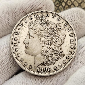 Puede incluir: Una moneda de plata Morgan de un dólar con el año 1895 inscrito en la parte inferior. La moneda presenta un perfil de Lady Liberty con el pelo suelto y una corona de hojas de laurel. La inscripción "E PLURIBUS UNUM" es visible alrededor del borde de la moneda.