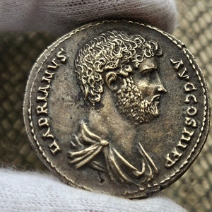 Puede incluir: Moneda romana de plata con un retrato de un hombre barbudo con el pelo rizado. La inscripción alrededor del borde dice "IMP COS VII AVG PONT MAX TR P XII".