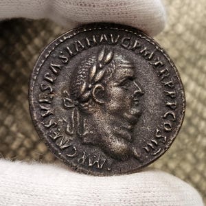 Puede incluir: Moneda romana de plata con un retrato de un hombre con una corona de laurel. La inscripción alrededor del retrato dice "IMP CAES L FLAVIUS AVG GRATRP COS III".