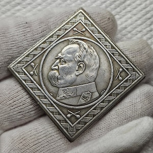 May include: A silver-toned, square-shaped commemorative medal with a raised image of a man's profile. The medal has a decorative border with a repeating pattern of crossed sticks. The year 1931 is inscribed on the medal.