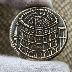 Puede incluir: Una moneda de plata con un diseño detallado de un edificio con un techo arqueado y columnas. La moneda tiene un borde elevado y una superficie lisa.