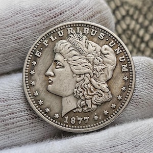 Puede incluir: Una moneda de plata Morgan de un dólar con el año 1877 inscrito en la parte inferior. La moneda presenta un perfil de Lady Liberty con la inscripción "E. PLURIBUS UNUM" alrededor del borde.