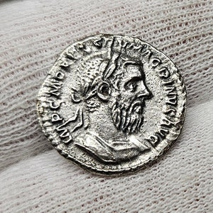 Puede incluir: Una moneda romana de plata que muestra el perfil de un hombre con barba y corona de laurel. La moneda tiene inscripciones detalladas en los bordes, lo que demuestra su importancia histórica. La superficie de la moneda muestra signos de desgaste.