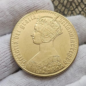 Puede incluir: Una moneda de oro con el perfil de una figura coronada, con la inscripción "Victoria Dei Gratia Britannia Regina". La moneda tiene detalles intrincados y un acabado pulido.