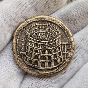 Puede incluir: Una moneda de color bronce con un relieve detallado de un anfiteatro romano. El diseño de la moneda incluye aberturas arqueadas y asientos escalonados, con una apariencia desgastada. La moneda es redonda y parece ser de metal, con un diámetro de unos 3 cm.