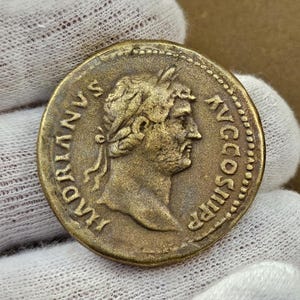 Puede incluir: Una moneda romana de bronce con el perfil de un hombre con una corona de laurel. La moneda tiene inscripciones en los bordes. La moneda está sujeta por una mano enguantada.