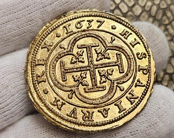 May include: A gold coin with intricate details, featuring a central cross design and the year 1637. The coin's edge is textured, and the surface displays engraved text. The coin is held in a gloved hand, showcasing its historical and collectible nature.