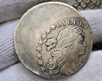 Może przedstawiać: Srebrna moneta z napisem "LIBERT" wygrawerowanym wokół krawędzi. Na monecie widnieje profil kobiety z rozpuszczonymi włosami i wstążką zawiązaną wokół głowy. Wokół głowy kobiety znajduje się 13 gwiazd.