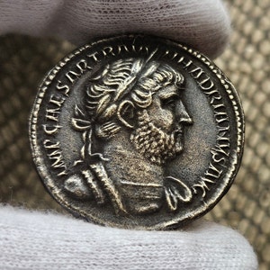 Puede incluir: Moneda romana de plata con un retrato del emperador Adriano en un lado y la inscripción "IMP CAESAR TRAIAN HADRIANVS AVG" en el otro.