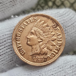 May include: An 1877 United States Indian Head penny coin, made of copper, with the words "United States of America" and the year "1877" inscribed around the edge.