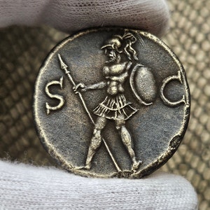 Puede incluir: Una moneda romana de plata con la imagen de un soldado sosteniendo una lanza y un escudo. La moneda tiene las letras "S" y "C" a cada lado del soldado.