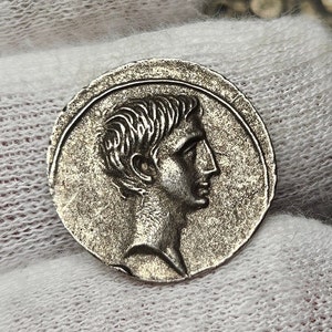 Puede incluir: Una moneda de plata que presenta el perfil de un hombre con detalles capilares y faciales. La moneda tiene forma circular con un borde texturizado y se encuentra sobre un fondo blanco.