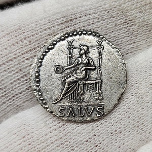 Puede incluir: Moneda de plata con un borde perlado, que presenta una figura sentada sosteniendo un objeto circular. La inscripción "SALVS" es visible debajo de la figura.