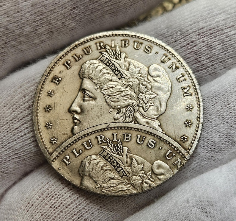 May include: A silver dollar coin with the words "E Pluribus Unum" and "Liberty" on it. The coin features a profile of a woman with a crown of leaves on her head.