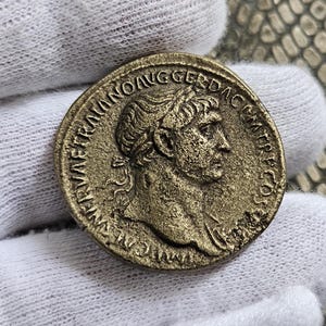 Puede incluir: Una moneda romana de bronce con la inscripción "IMP CAESAR AVG GER DAC IMP TR P COS", que representa un perfil de un hombre con una corona de laurel.
