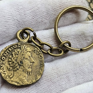 Puede incluir: Llavero de color latón antiguo con un colgante de moneda redondo. La moneda representa un perfil de una persona y tiene texto alrededor del borde. El llavero incluye una cadena y un anillo grande para llaves.