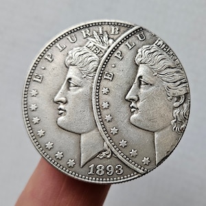 Może przedstawiać: Srebrna moneta dolara z dwoma portretami osób po obu stronach. Na monecie widnieją słowa "E Pluribus Unum" i "Liberty". Data "1893" jest nadrukowana na dole monety.