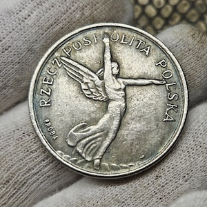 May include: A silver coin featuring a winged figure with outstretched arms, with the words "RZECZ POSPOLITA POLSKA" and "PROBA" around the edge. The coin has a detailed design and a slightly aged appearance.