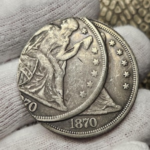May include: A silver dollar coin with the year 1870 on the front and the year 1870 on the back. The coin features a woman holding a torch and a shield.