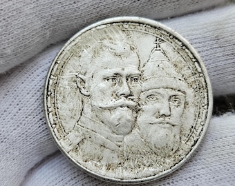 Könnte beinhalten: Eine kreisförmige silberfarbene Münze mit den geprägten Profilen zweier Männer mit Schnurrbärten und Bärten. Die Figur links trägt eine militärische Uniform, während die Figur rechts eine traditionelle spitze Kopfbedeckung mit Kreuz trägt.
