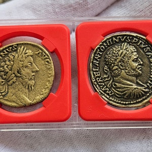 Puede incluir: Dos monedas romanas antiguas en vitrinas de plástico rojo. Las monedas son de bronce y presentan un perfil de un hombre con una corona de laurel en la cabeza. La inscripción en las monedas dice "IMP ANTONINVS PIVS AVG".