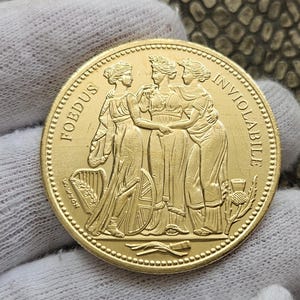 Puede incluir: Una moneda de oro con tres mujeres de pie juntas. La moneda tiene las palabras "Foedus Inviolable" inscritas alrededor del borde.