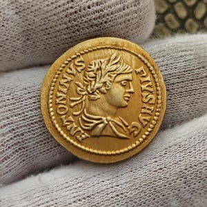 Puede incluir: Una moneda de oro con el retrato de una persona con una corona de laurel en la cabeza. La inscripción alrededor del retrato dice "IMP ANTONINVS PIVS AVG".