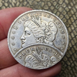 May include: A silver US dollar coin with the words "E Pluribus Unum" and "Liberty" on the obverse side. The coin features a portrait of a woman with flowing hair and a wreath on her head.