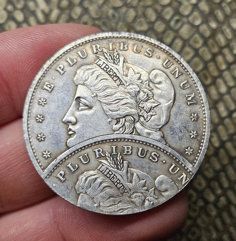May include: A silver US dollar coin with the words "E Pluribus Unum" and "Liberty" on the obverse side. The coin features a portrait of a woman with flowing hair and a wreath on her head.