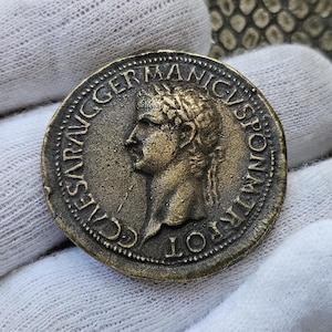 Puede incluir: Una moneda romana de bronce con la inscripción "CAESAR AVG GERM" y un retrato de un hombre con una corona de laurel.
