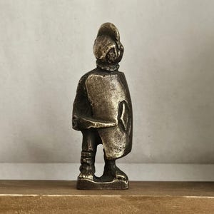 Puede incluir: Figura de color bronce de una persona con vestimenta histórica, posiblemente un soldado o noble. La figura está de pie, con un abrigo largo y un sombrero. La estatua mide aproximadamente 10 cm de altura.