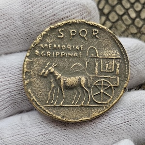 Puede incluir: Una moneda romana de bronce con la inscripción "SPQR MEMORIAE AGRIPPINAE" y la representación de un carro tirado por dos caballos.