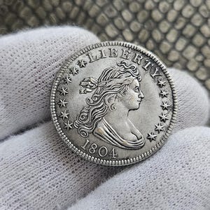 Puede incluir: Una moneda de tono plateado que presenta el retrato de perfil de una figura mirando hacia la derecha, con la palabra LIBERTY arqueada arriba y el año 1804 inscrito abajo. La moneda está rodeada por trece estrellas y un borde estriado.
