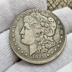 May include: A silver US dollar coin with the inscription "E Pluribus Unum" and the year "1921" on the back. The coin features a profile of a woman with flowing hair and a wreath on her head.