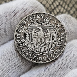 Puede incluir: Una moneda de plata de medio dólar de los Estados Unidos. La moneda presenta un águila con las alas extendidas, las palabras "UNITED STATES OF AMERICA", "IN GOD WE TRUST" y "HALF DOLLAR". La moneda se sostiene en un guante blanco.