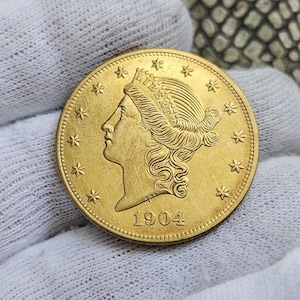 May include: A gold coin featuring a profile of a woman with the word "LIBERTY" above her head. The coin is dated 1904 and has stars around the edge. The coin is held against a white fabric background.