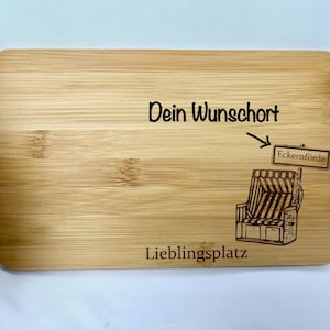 Puede incluir: Tabla de cortar rectangular de bambú con texto grabado y una ilustración de una silla de playa. El texto incluye "Dein Wunschort" y "Lieblingsplatz", con una flecha que apunta a un letrero que dice "Eckernförde".