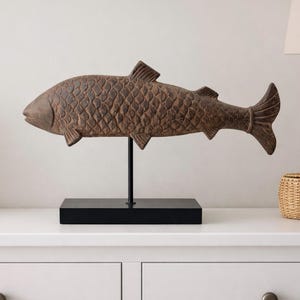 Può includere: Scultura di pesce in legno marrone con dettagli di squame e pinne. Il pesce è montato su un supporto e una base in metallo nero. Il pezzo decorativo è esposto su una superficie bianca, suggerendo un tema di arredamento per la casa.