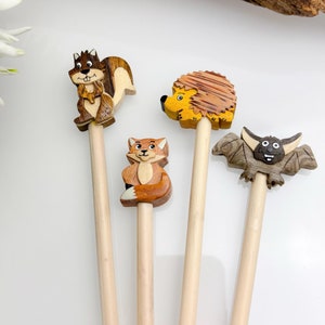 Op de afbeelding: Vier houten potloden met dierenfiguren: een eekhoorn, een vos, een egel en een vleermuis. De eekhoorn is bruin en bruin, de vos is oranje en wit, de egel is geel en bruin en de vleermuis is grijs. De potloden liggen op een witte ondergrond.