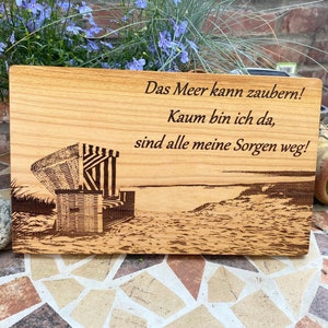 Może przedstawiać: Prostokątna drewniana tablica z wygrawerowaną sceną plażową i niemieckim tekstem: "Das Meer kann zaubern! Kaum bin ich da, sind alle meine Sorgen weg!" Przedstawia leżak plażowy, linię brzegową i ocean. Tablica jest z jasnego drewna.