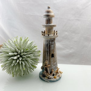 May include: A decorative wooden lighthouse with a distressed, weathered finish. The lighthouse features a light brown and white color scheme, with an anchor and seashells at the base. A green and white decorative plant is to the left.