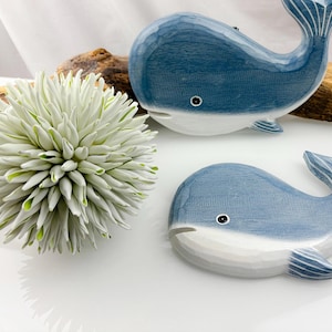 May include: Two blue and white whale figurines. The whales have a carved wood appearance with a light blue body and white belly. The tail fins have white carved lines. The whales are decorative items.