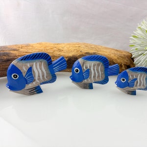 May include: Three wooden fish ornaments with blue and grey details. The fish have blue heads and tails, grey bodies with white stripes, and white eyes. They are arranged in a row on a white surface, with a piece of driftwood in the background.